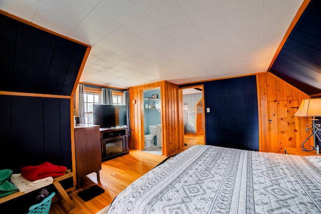 bedroom with ensuite bathroom, lofted ceiling, wood walls, and light hardwood / wood-style floors