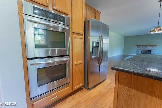 kitchen with a stone fireplace, decorative light fixtures, stainless steel appliances, dark stone countertops, and light hardwood / wood-style floors