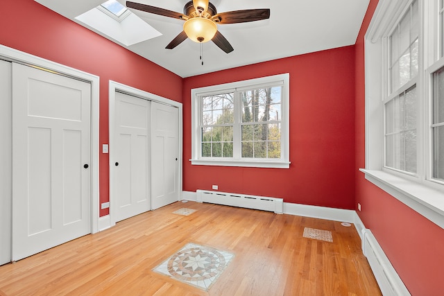 unfurnished bedroom with light wood-type flooring, a skylight, ceiling fan, and a baseboard heating unit