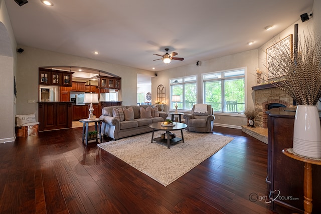 living room featuring ceiling fan, a stone fireplace, and dark wood-type flooring