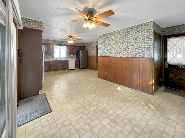 unfurnished living room with wallpapered walls, a wainscoted wall, a ceiling fan, and wooden walls