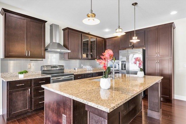 kitchen with wall chimney range hood, a kitchen island with sink, decorative light fixtures, stainless steel appliances, and dark hardwood / wood-style floors