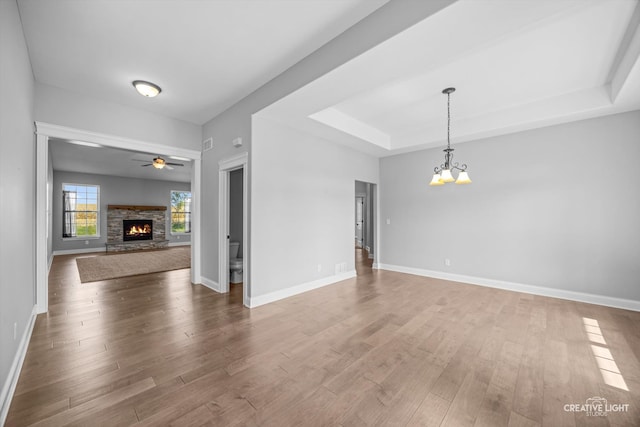 unfurnished living room featuring a fireplace, ceiling fan with notable chandelier, a tray ceiling, and hardwood / wood-style flooring