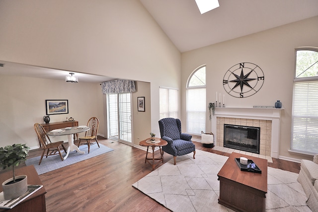 living room featuring high vaulted ceiling, light hardwood / wood-style flooring, a skylight, and a tile fireplace