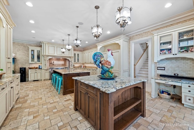 kitchen featuring tasteful backsplash, cream cabinets, decorative light fixtures, a kitchen island with sink, and crown molding