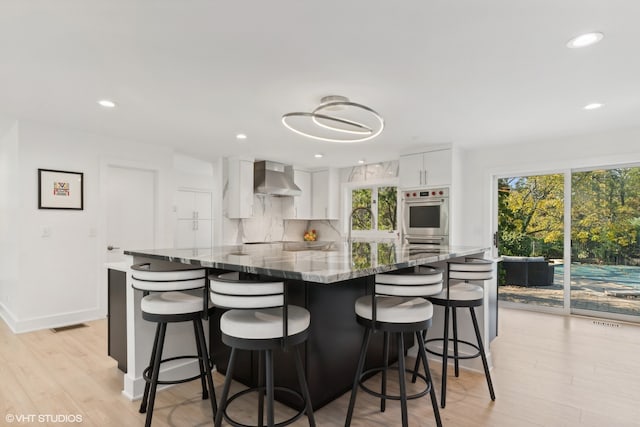 kitchen with stone countertops, wall chimney range hood, a breakfast bar area, light wood-type flooring, and white cabinetry