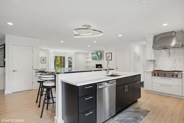 kitchen with wall chimney range hood, white cabinetry, stainless steel appliances, and an island with sink