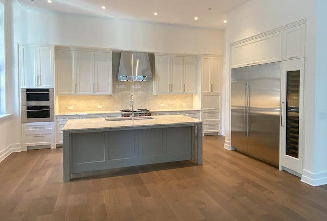 kitchen with a kitchen island with sink, wall chimney exhaust hood, white cabinetry, and built in refrigerator