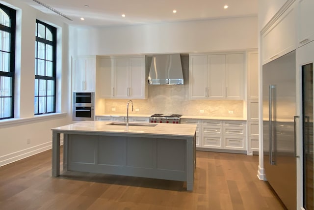 kitchen with wall chimney range hood, sink, an island with sink, appliances with stainless steel finishes, and white cabinetry