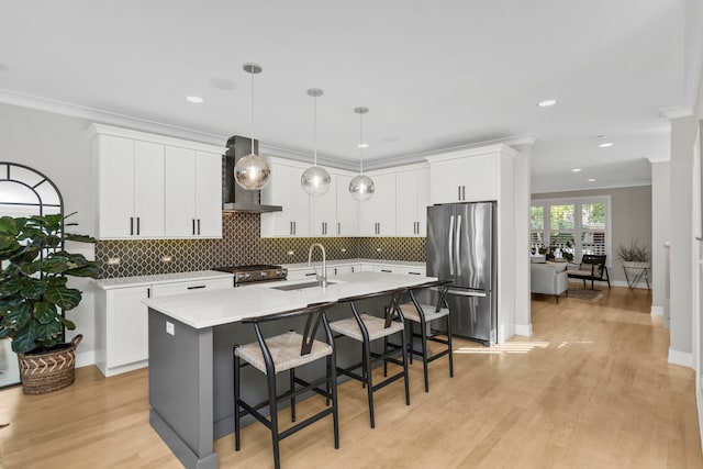 kitchen featuring an island with sink, white cabinets, sink, wall chimney range hood, and appliances with stainless steel finishes