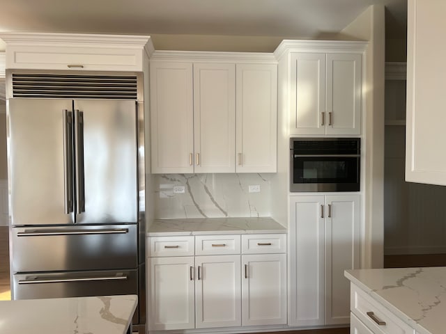 kitchen featuring built in refrigerator, white cabinets, oven, backsplash, and light stone countertops