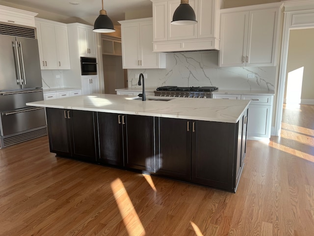 kitchen with stainless steel appliances, a center island with sink, backsplash, hanging light fixtures, and light hardwood / wood-style flooring