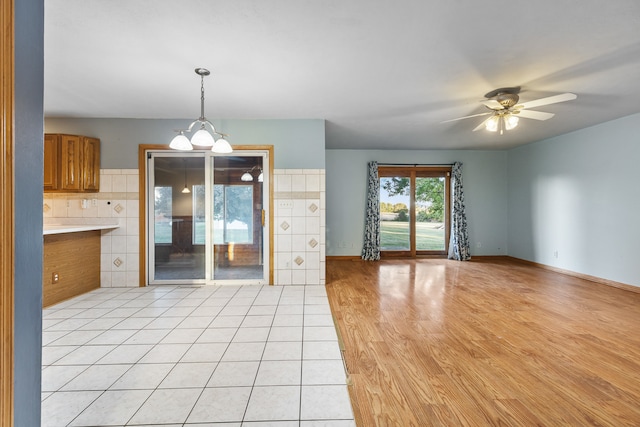 unfurnished living room with tile walls, ceiling fan with notable chandelier, and light wood-type flooring