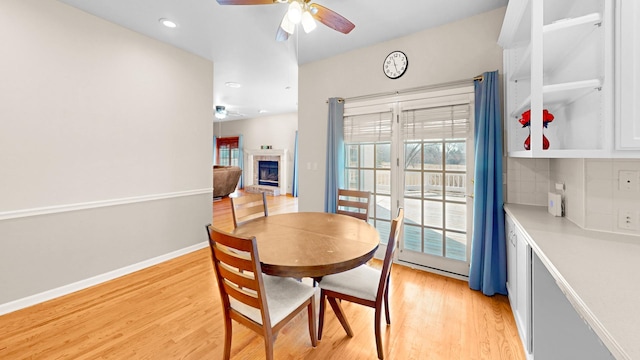 dining room featuring a ceiling fan, a fireplace with raised hearth, light wood-style floors, and baseboards