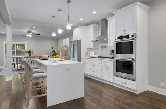 kitchen with stainless steel appliances, wall chimney range hood, pendant lighting, a center island with sink, and white cabinets