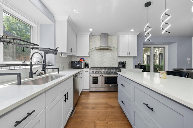 kitchen with appliances with stainless steel finishes, hanging light fixtures, wall chimney exhaust hood, white cabinets, and hardwood / wood-style flooring