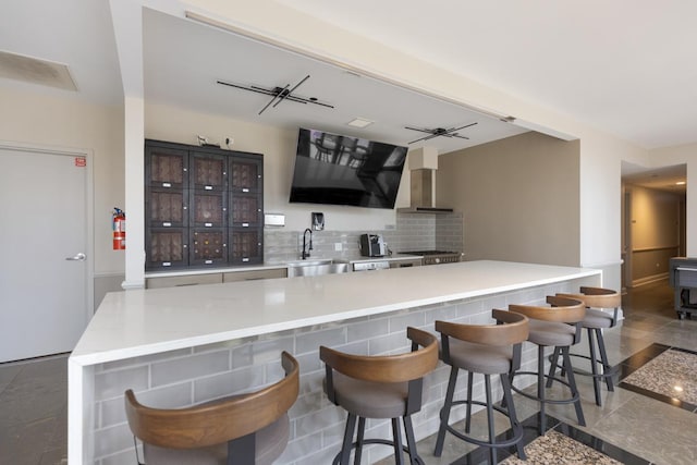 kitchen featuring decorative backsplash, dark brown cabinetry, wall chimney range hood, sink, and a breakfast bar area