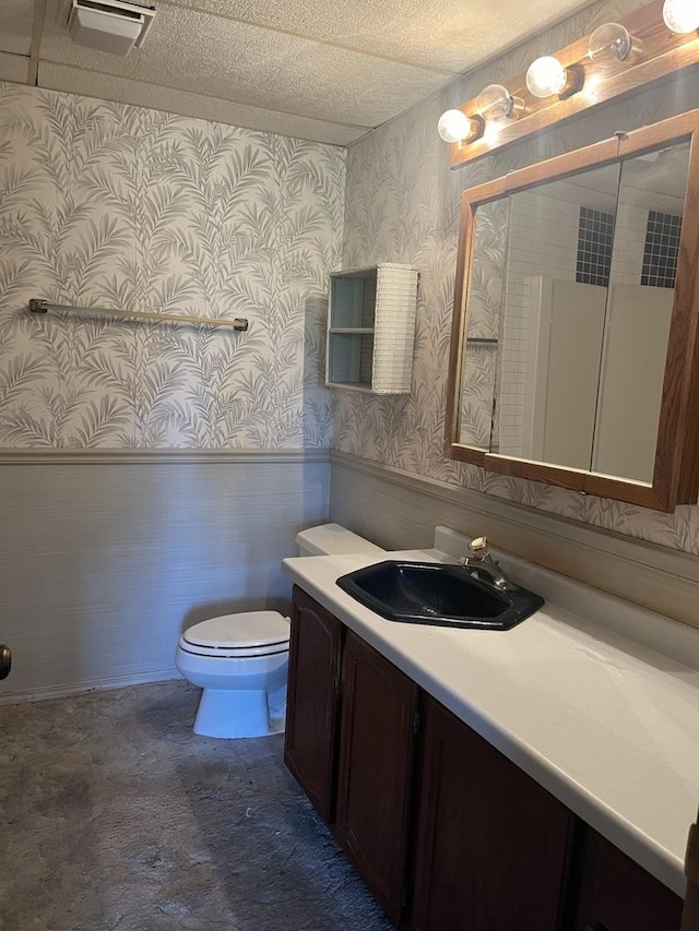 bathroom with wallpapered walls, toilet, vanity, wainscoting, and a textured ceiling