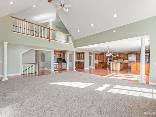 unfurnished living room with light carpet, high vaulted ceiling, ceiling fan with notable chandelier, and decorative columns