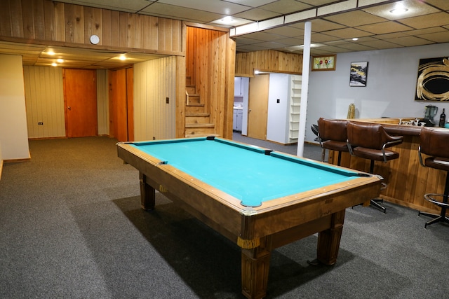 recreation room featuring a paneled ceiling, wooden walls, and dark carpet