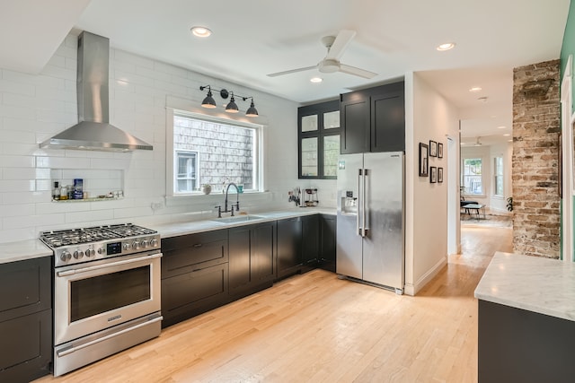 kitchen featuring premium appliances, wall chimney range hood, sink, light wood-type flooring, and tasteful backsplash