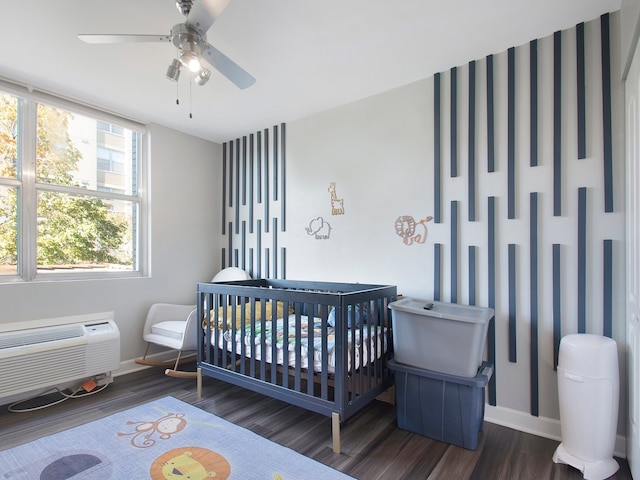 bedroom with dark wood-type flooring, ceiling fan, a wall unit AC, and a crib