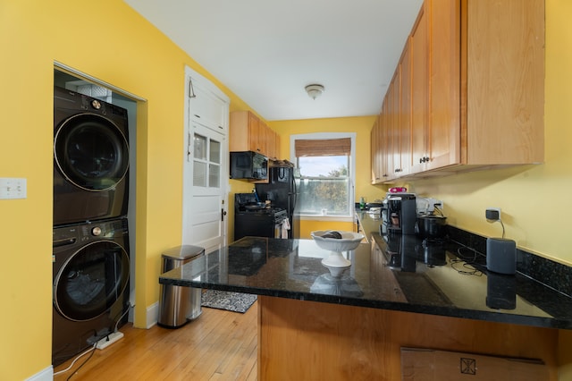 kitchen featuring black appliances, kitchen peninsula, dark stone counters, light hardwood / wood-style flooring, and stacked washer and clothes dryer