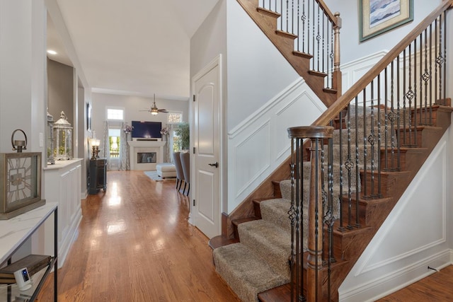 stairway featuring ceiling fan, a decorative wall, wood finished floors, and a glass covered fireplace