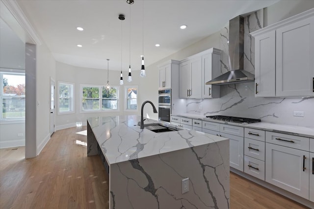 kitchen with sink, an island with sink, hanging light fixtures, stainless steel appliances, and wall chimney exhaust hood