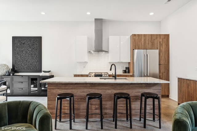 kitchen with wall chimney exhaust hood, high quality fridge, white cabinets, a breakfast bar, and a center island with sink