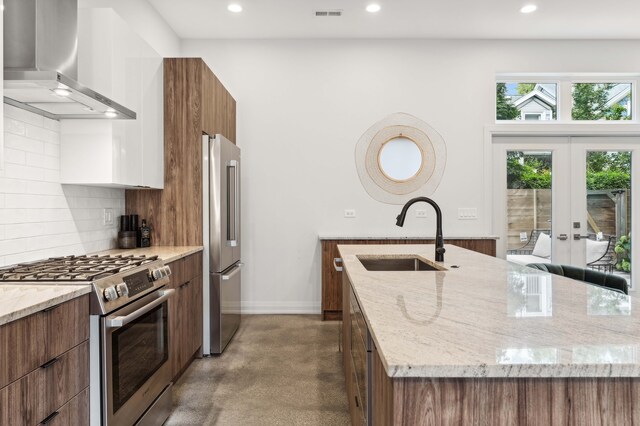 kitchen with wall chimney range hood, a center island with sink, sink, premium appliances, and light stone counters