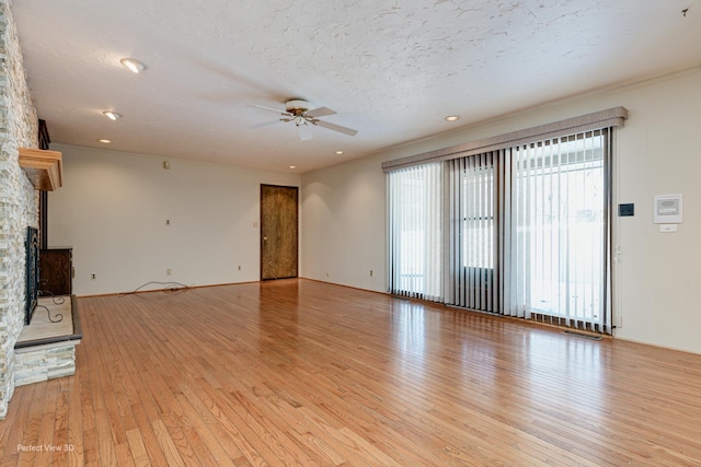 unfurnished living room featuring ceiling fan, light wood-type flooring, a stone fireplace, a textured ceiling, and ornamental molding