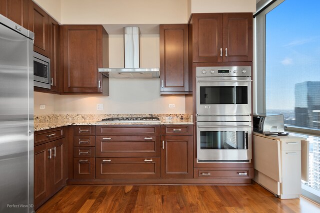 kitchen with appliances with stainless steel finishes, wall chimney exhaust hood, light stone countertops, and dark wood-type flooring