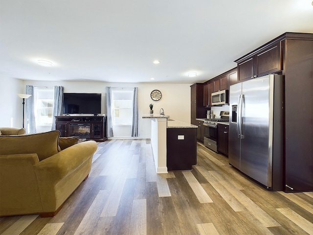 kitchen featuring appliances with stainless steel finishes, dark brown cabinets, light stone counters, light hardwood / wood-style flooring, and a center island with sink