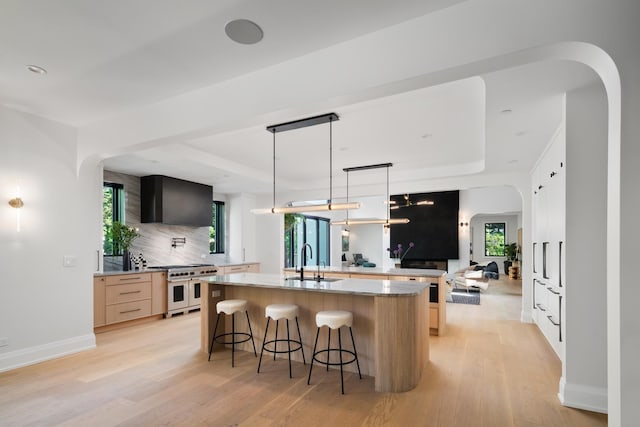 kitchen featuring range with two ovens, light wood-type flooring, a kitchen island with sink, and light brown cabinetry