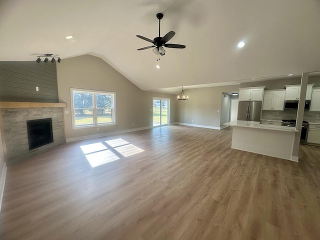 unfurnished living room featuring ceiling fan with notable chandelier, high vaulted ceiling, a stone fireplace, and light hardwood / wood-style floors