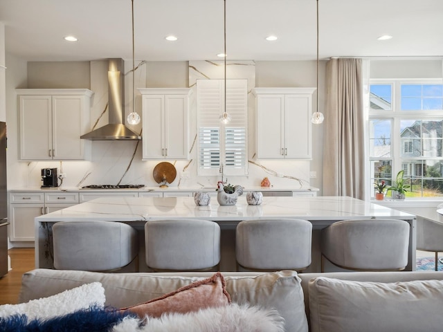 kitchen featuring light stone countertops, backsplash, wall chimney exhaust hood, pendant lighting, and white cabinetry