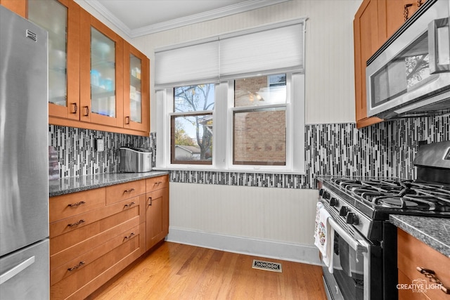 kitchen featuring crown molding, dark stone counters, stainless steel appliances, light wood-type flooring, and backsplash