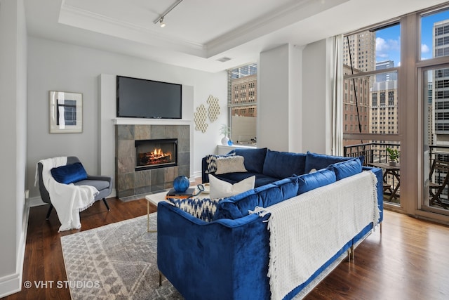 living room with a raised ceiling, dark hardwood / wood-style flooring, a tile fireplace, crown molding, and track lighting