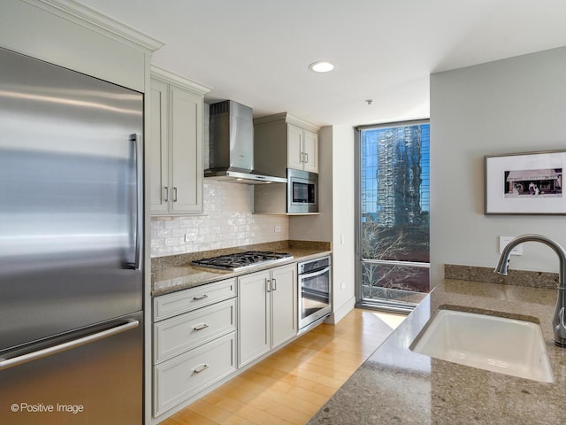 kitchen with dark stone counters, wall chimney range hood, sink, built in appliances, and light wood-type flooring