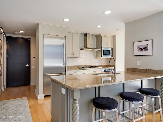 kitchen with sink, wall chimney exhaust hood, kitchen peninsula, built in appliances, and light hardwood / wood-style floors