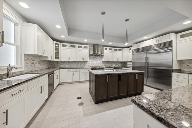 kitchen featuring stainless steel appliances, a raised ceiling, sink, wall chimney range hood, and an island with sink