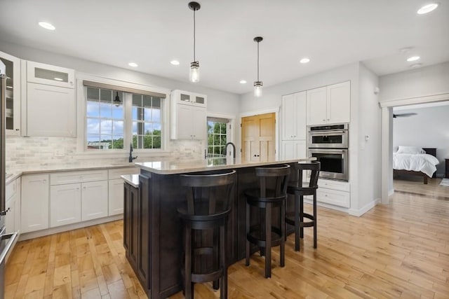 kitchen with double oven, a center island with sink, white cabinets, light hardwood / wood-style floors, and hanging light fixtures