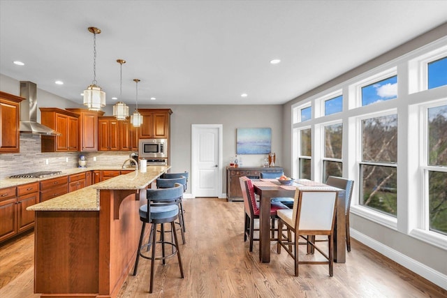 kitchen featuring wall chimney range hood, decorative light fixtures, a breakfast bar area, a center island with sink, and appliances with stainless steel finishes