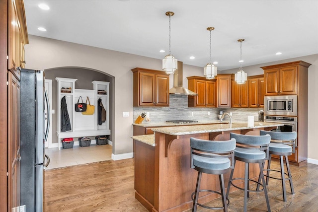 kitchen featuring a kitchen breakfast bar, stainless steel appliances, wall chimney range hood, hanging light fixtures, and an island with sink