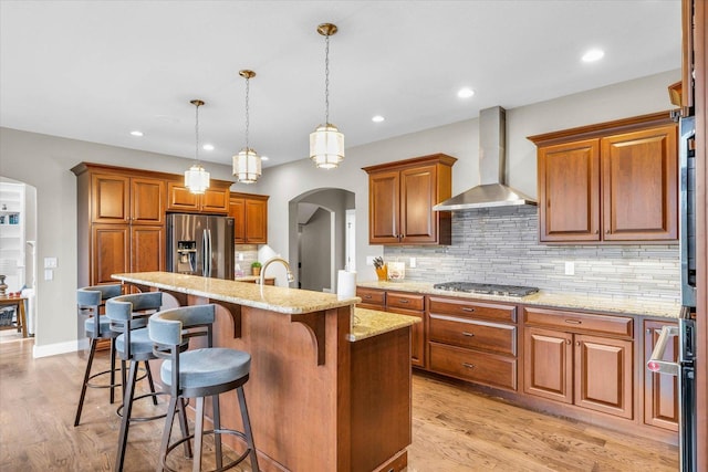 kitchen with a kitchen island with sink, wall chimney range hood, hanging light fixtures, decorative backsplash, and stainless steel appliances