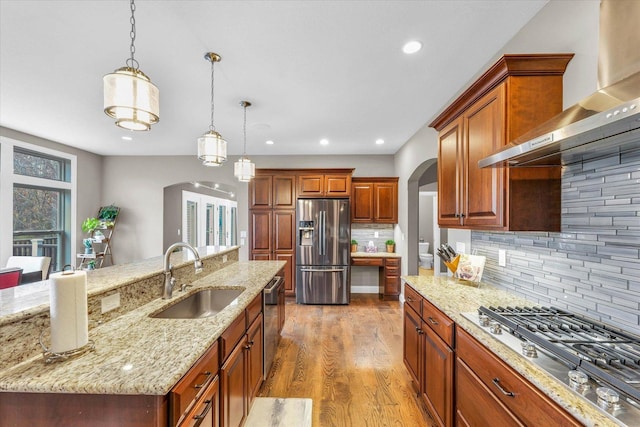 kitchen with appliances with stainless steel finishes, wall chimney exhaust hood, sink, a large island with sink, and decorative light fixtures