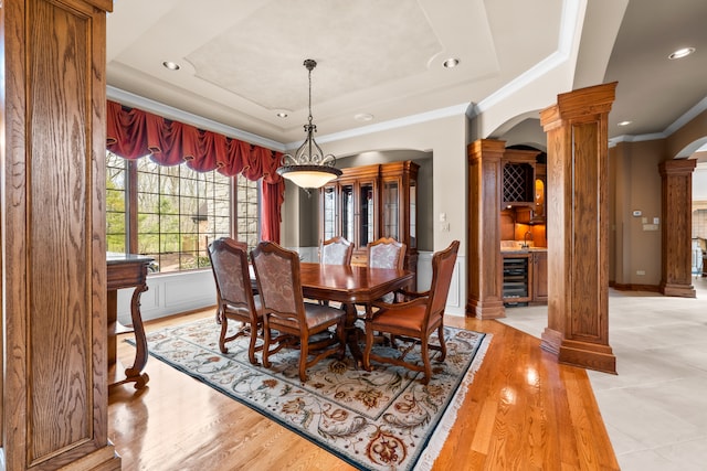 dining area with a raised ceiling, ornate columns, wine cooler, ornamental molding, and light hardwood / wood-style flooring