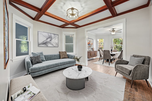 living room with beam ceiling, coffered ceiling, crown molding, wood-type flooring, and ceiling fan with notable chandelier