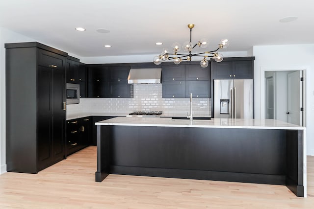 kitchen featuring sink, appliances with stainless steel finishes, an island with sink, light hardwood / wood-style flooring, and wall chimney range hood